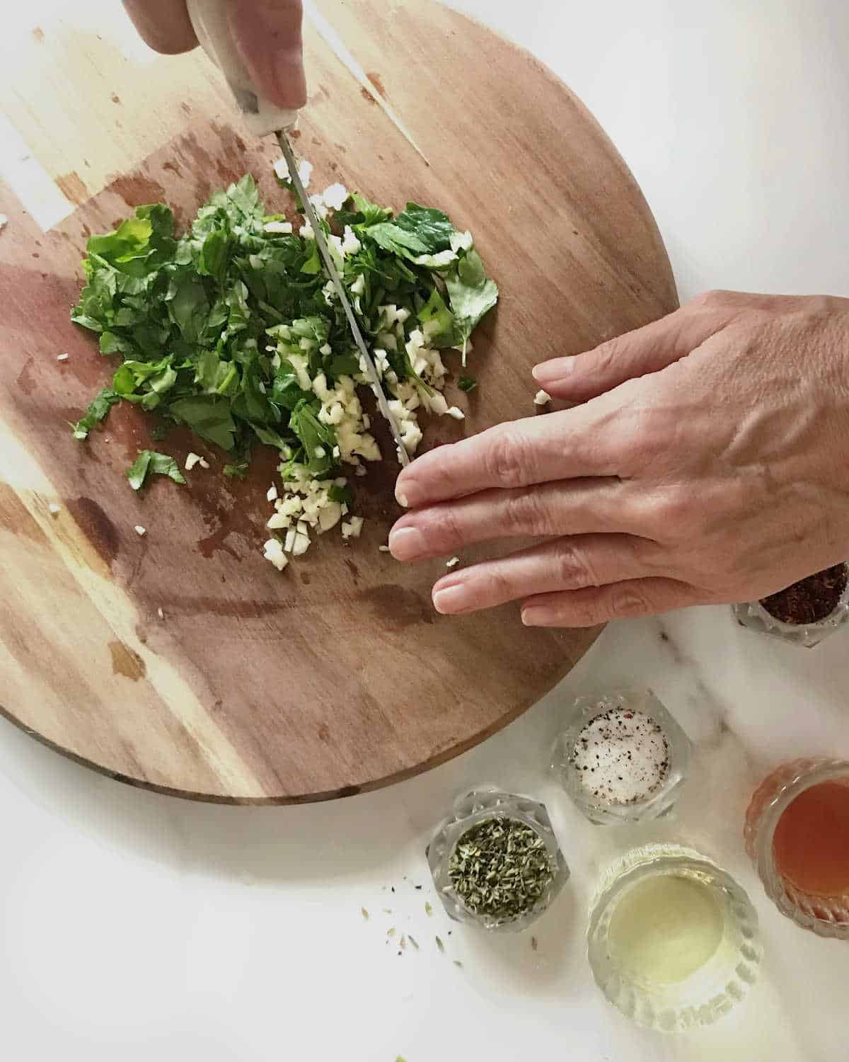 Hands chopping parsley and garlic on a wooden board, white surface with other ingredients in containers.
