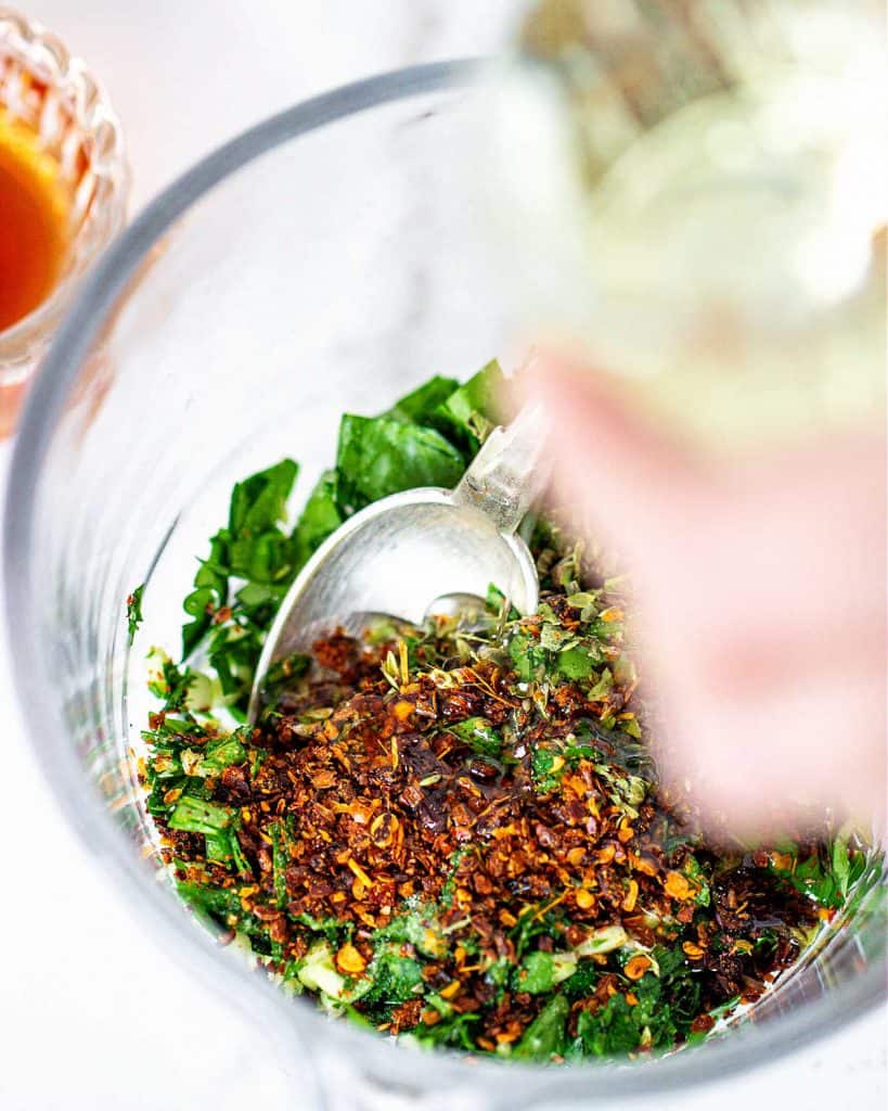 Glass jar with chopped parsley and red pepper flakes. Hand on top about to add oil.