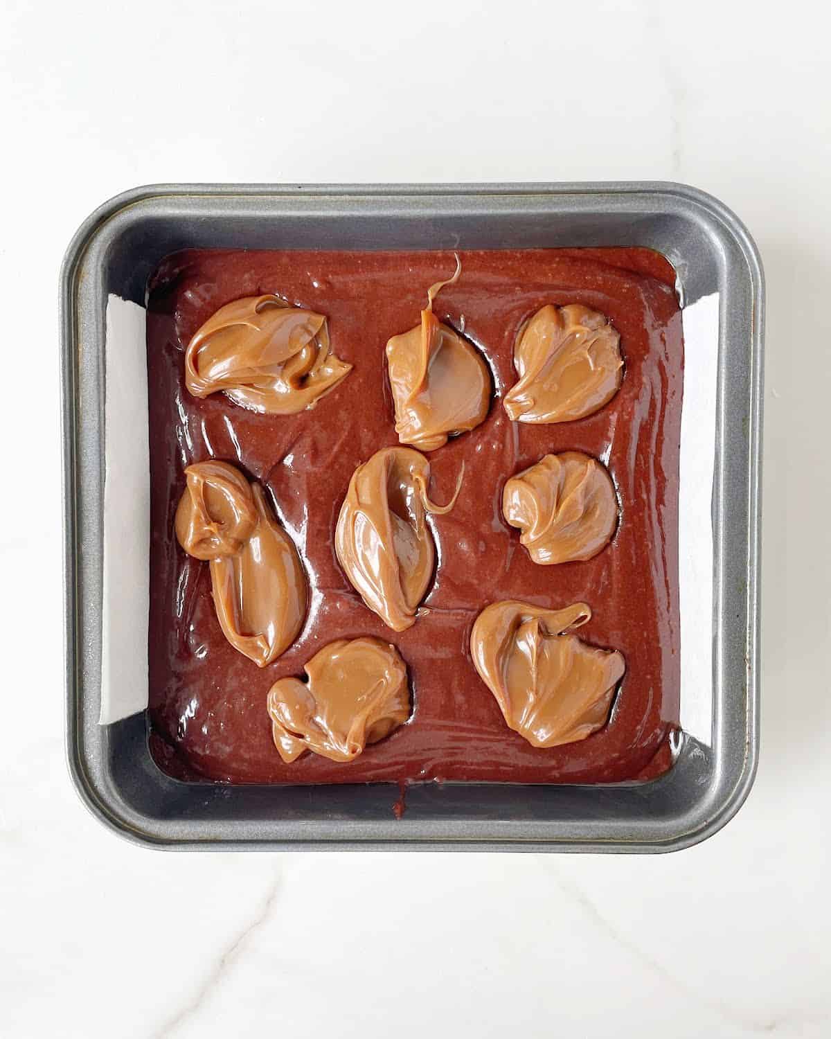 Dollops of dulce de leche on brownie batter in a dark metal square pan on a white marble surface. 