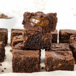Pile and rows of brownies oozing dulce de leche on parchment paper with a white background.