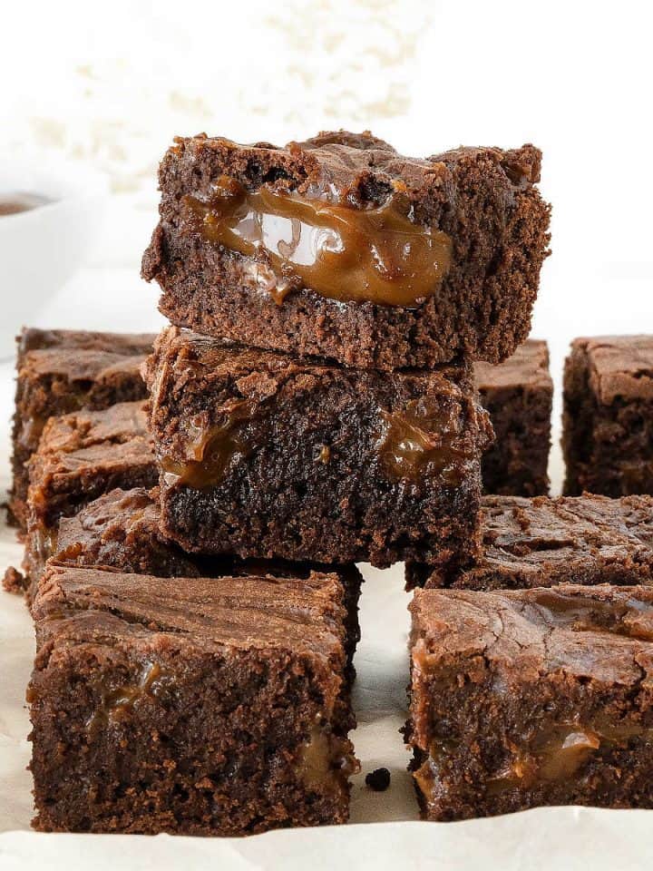 Pile and rows of brownies oozing dulce de leche on parchment paper with a white background.