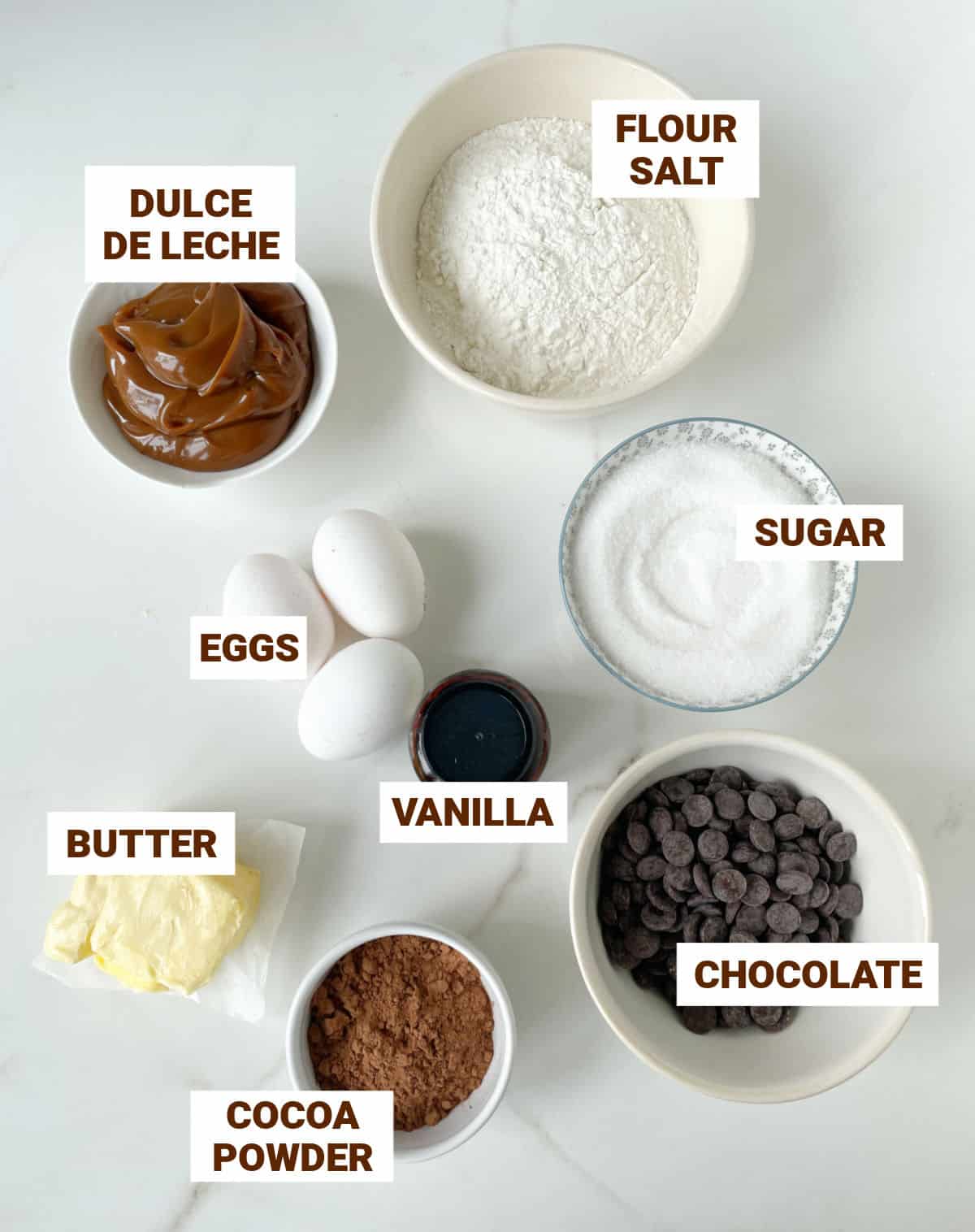 Bowls containing ingredients for dulce de leche brownies on a white surface including butter, flour, cocoa powder, vanilla, eggs, sugar.