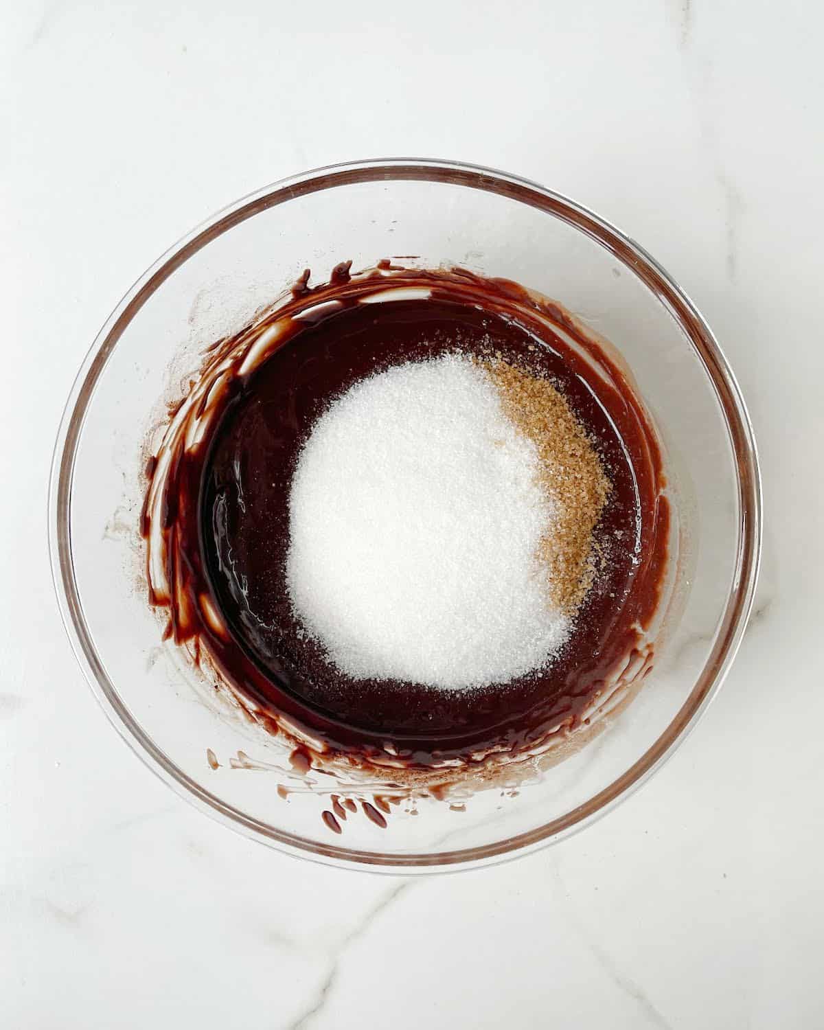 Sugars added to brownie batter in a glass bowl. White marble surface.