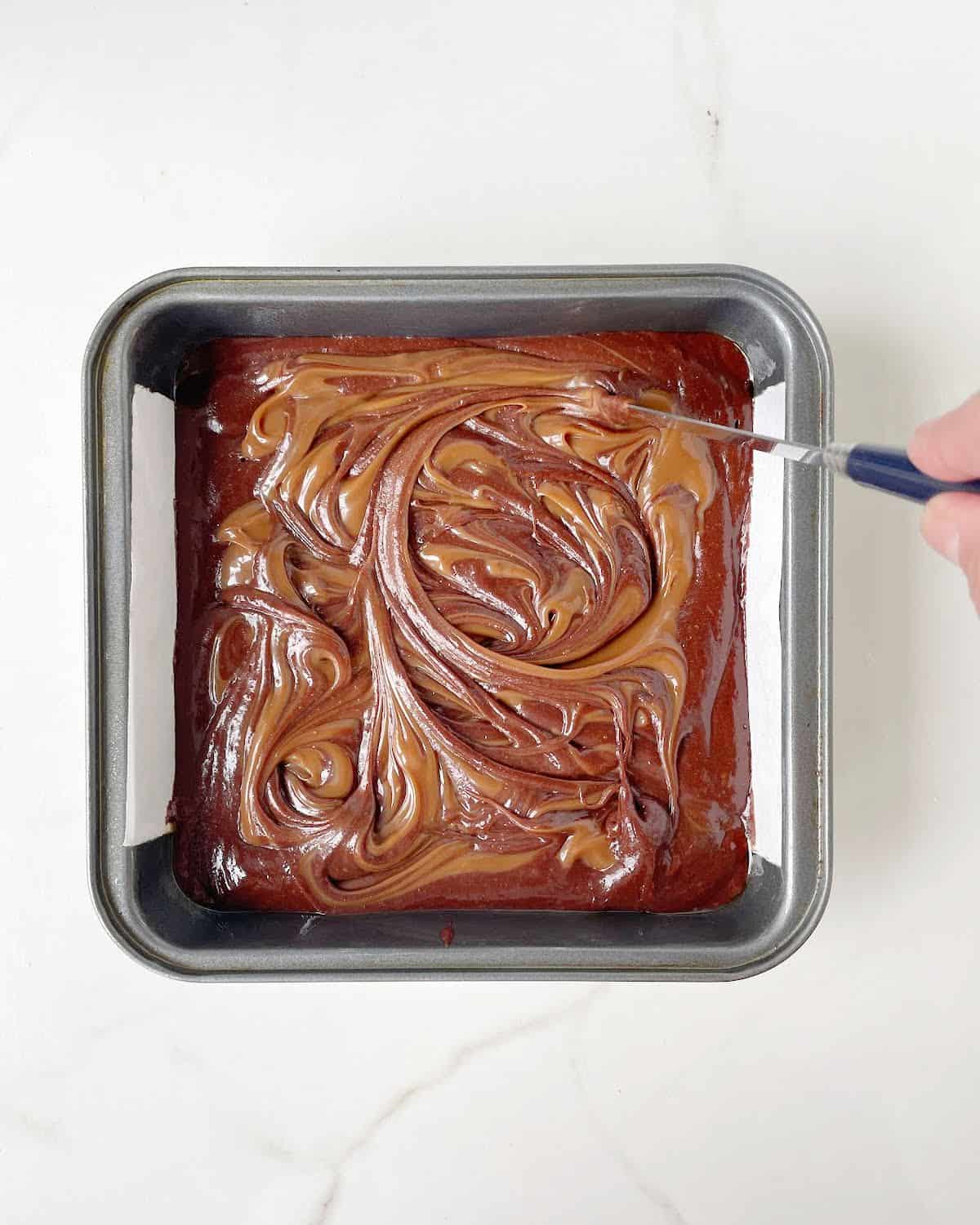 Knife swirling dulce de lech on top of brownie batter in a square metal pan on a white surface. 