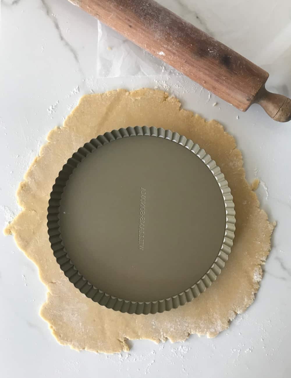 Round of sweet dough with pie pan on top, white surface, rolling pin.