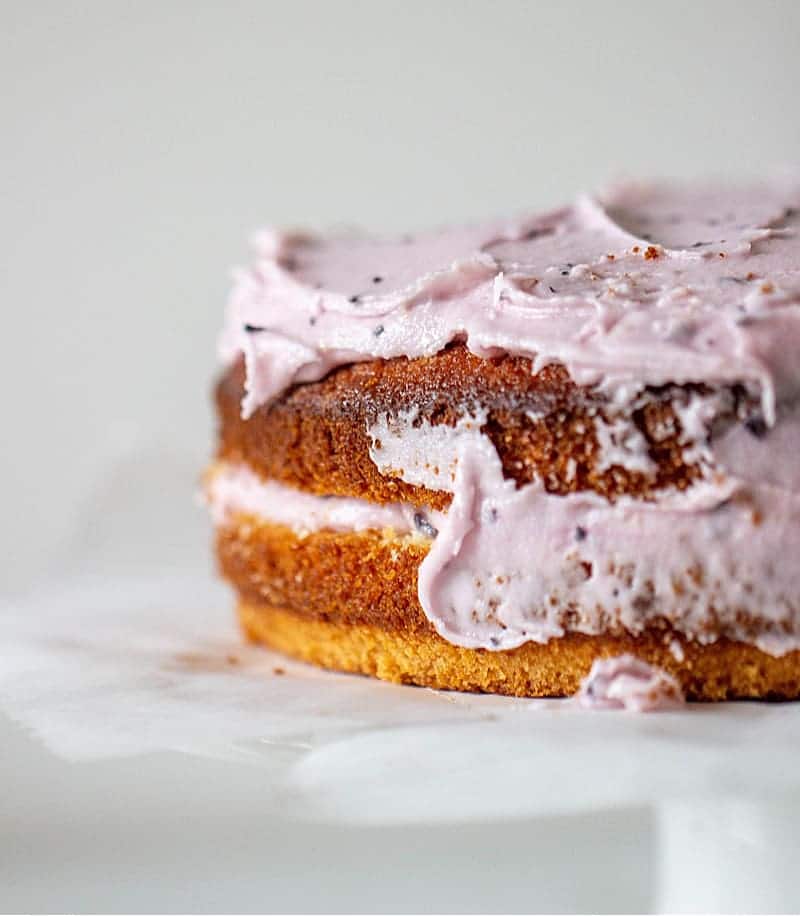 Partially frosted lemon blueberry cake on a white cake stand.