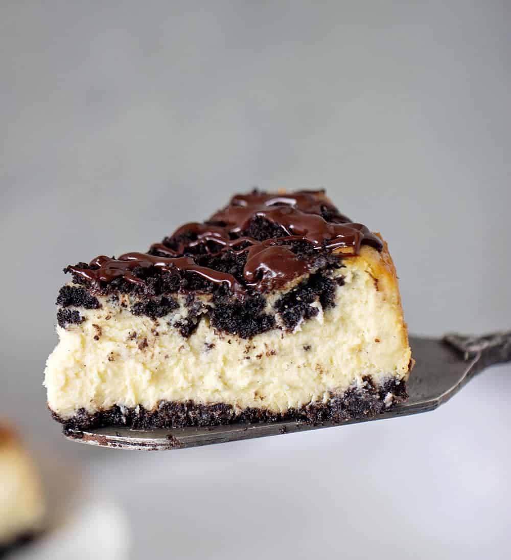 Slice of Oreo Cheesecake with chocolate topping on cake server, grey background