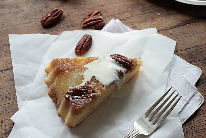 Slice of pear upside down cake on white parchment paper, wooden table, scattered pecans.