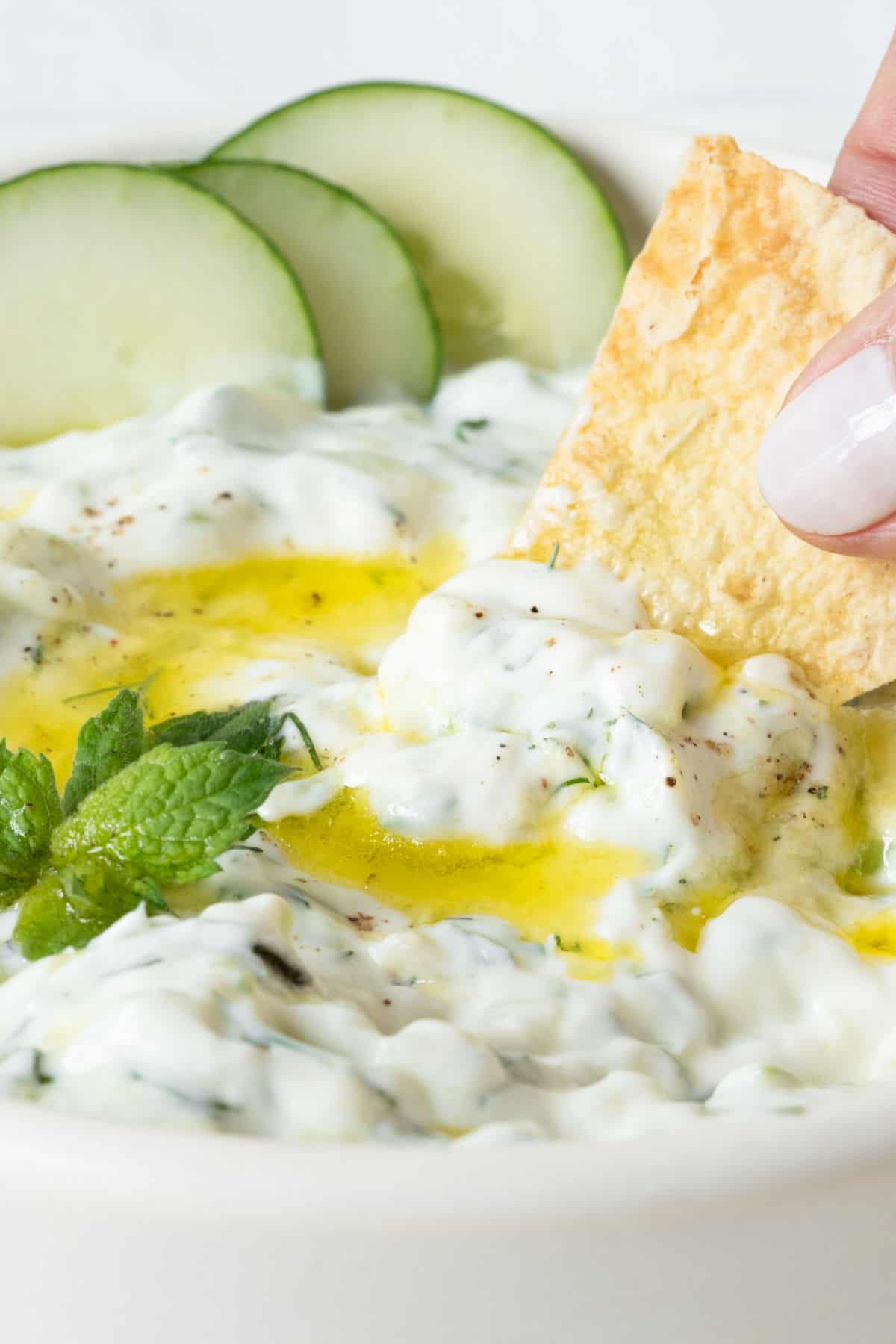 Close up of pita chip inside a white bowl with tzatziki sauce. Cucumber rounds, herbs, and olive oil. 