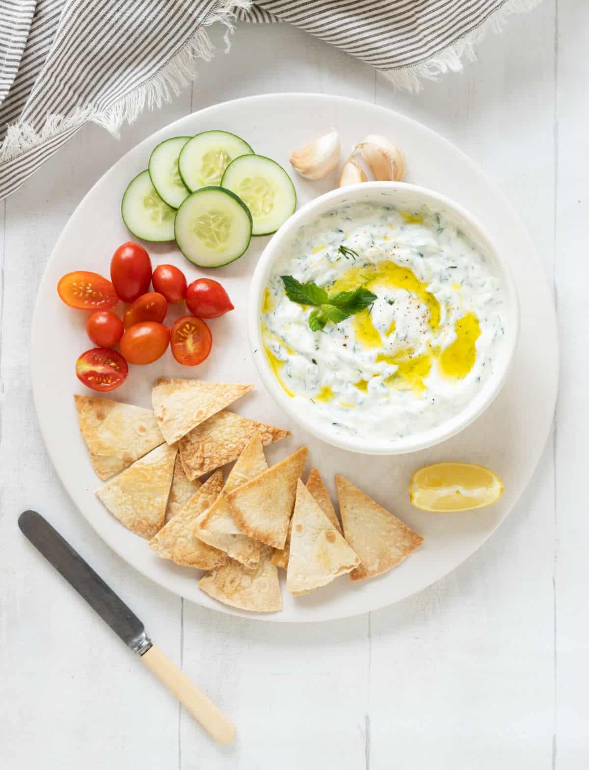 White platter with bowl of tzatziki, cucumber slices, cherry tomatoes, pita chips. White surface, striped cloth. 