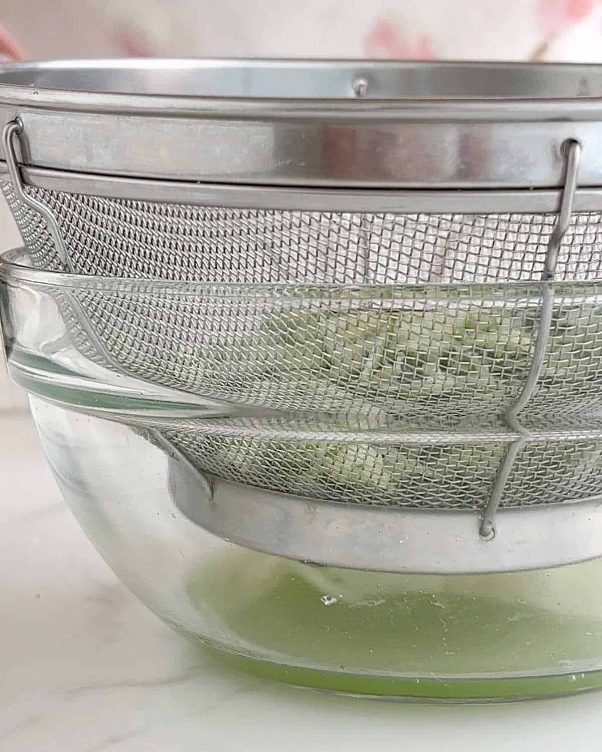 Grated cucumber draining in a metal colander set on a glass bowl.