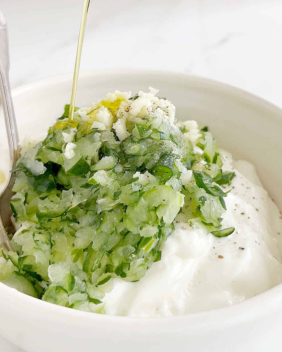 Drizzling olive oil over plain yogurt with grated cucumber in a white bowl. 