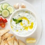 Tzatziki sauce in a white bowl on a white platter with cucumber slices, pita chips, and cherry tomatoes.