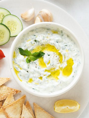 Tzatziki sauce in a white bowl on a white platter with cucumber slices, pita chips, and cherry tomatoes.
