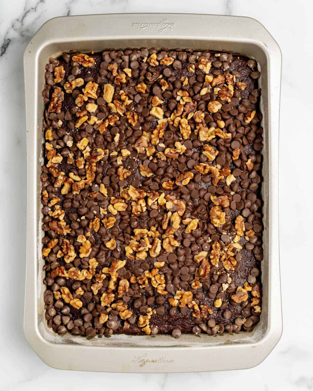 Metal rectangular cake pan with walnut and chocolate chip topped cake. White marble surface.