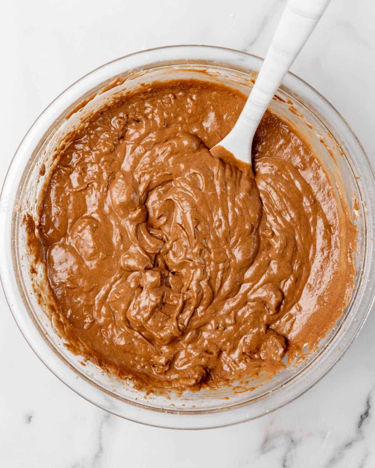 Chocolate date cake batter in a glass bowl with a spatula on a marble surface.