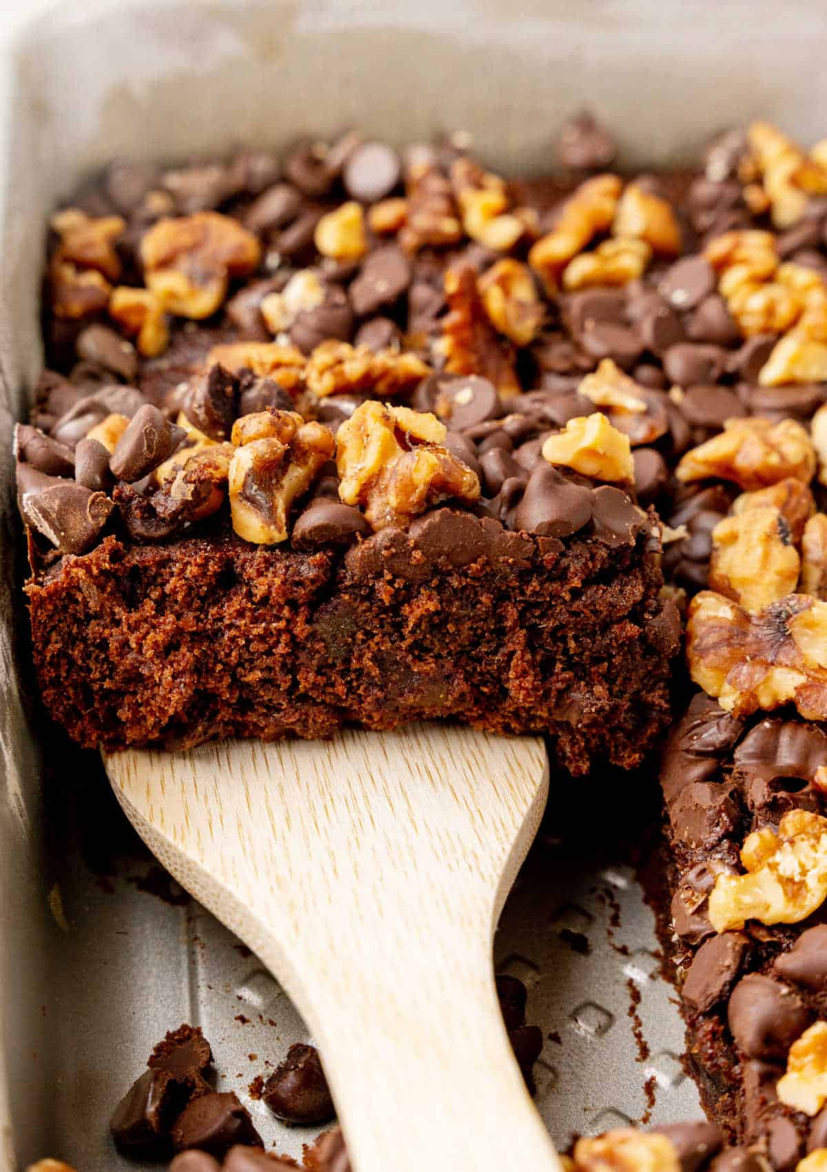 Wooden spatula lifting piece of date cake with chocolate chips and walnuts from the pan. 