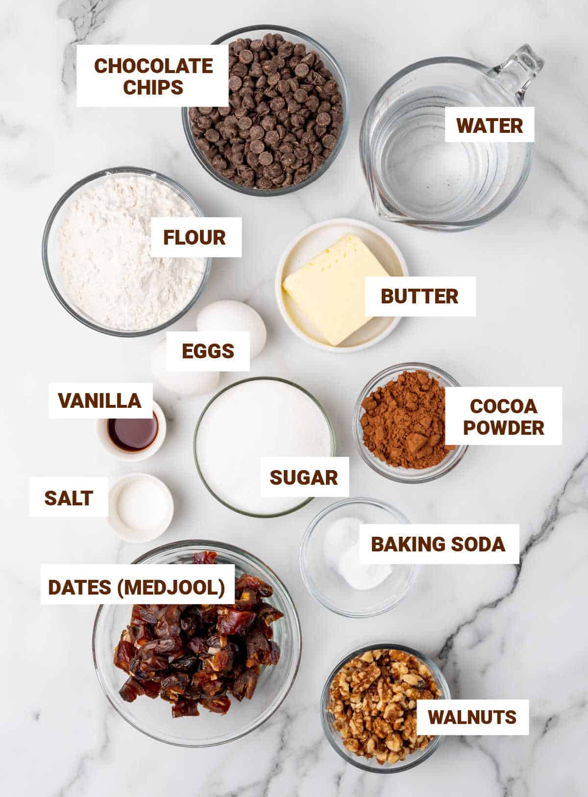 Bowls on white marble surface containing ingredients for chocolate date cake including walnuts, flour mixture, butter, eggs, sugar, cocoa powder, vanilla.