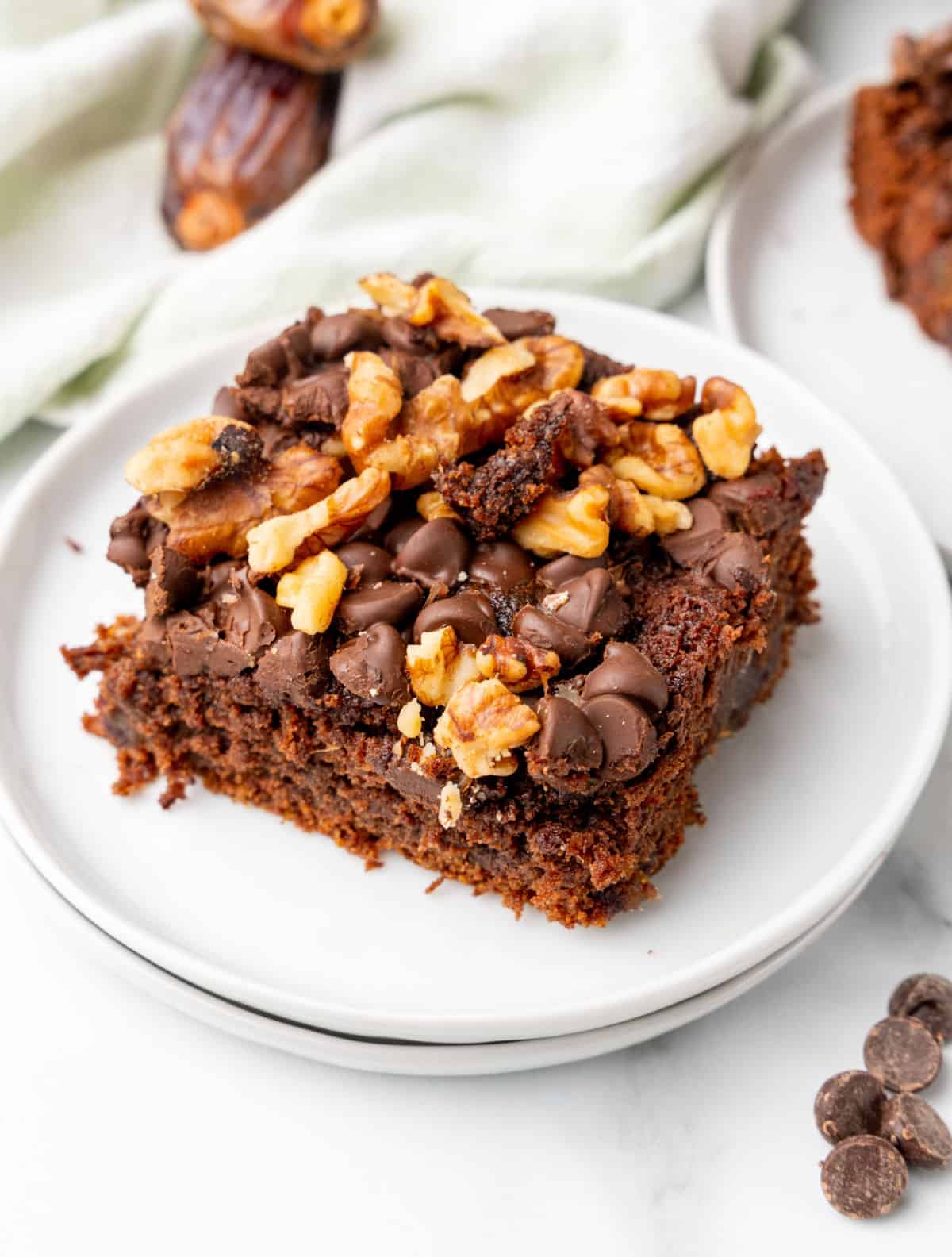 Square of chocolate chip walnut date cake on stack of two white plates. White surface, kitchen towel, loose dates around. 