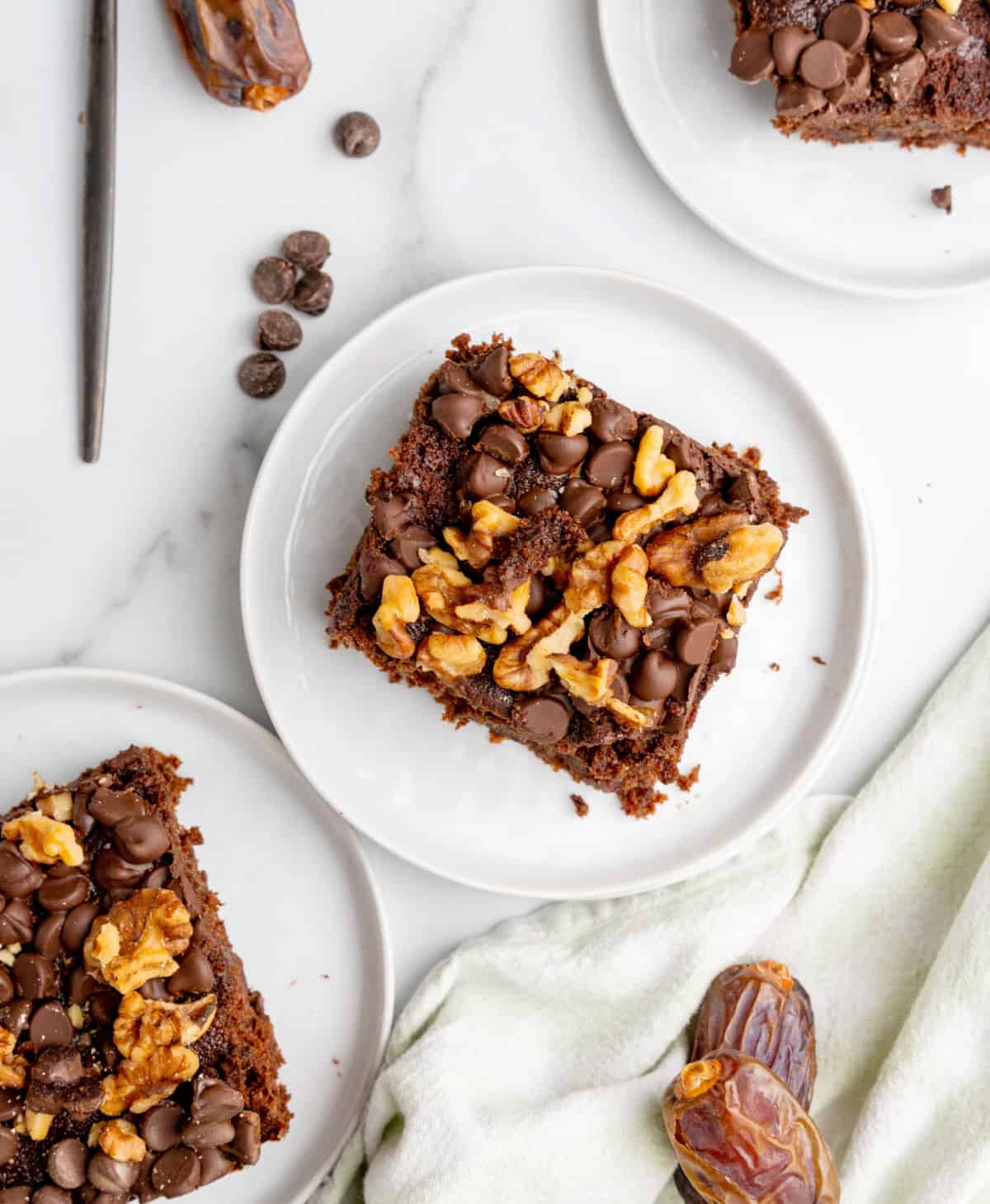 Top view of white plates with squares of walnut chocolate chip topped date cake on a white surface.