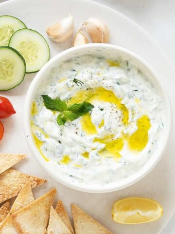 Tzatziki sauce in a white bowl on a white platter with cucumber slices, pita chips, and cherry tomatoes.