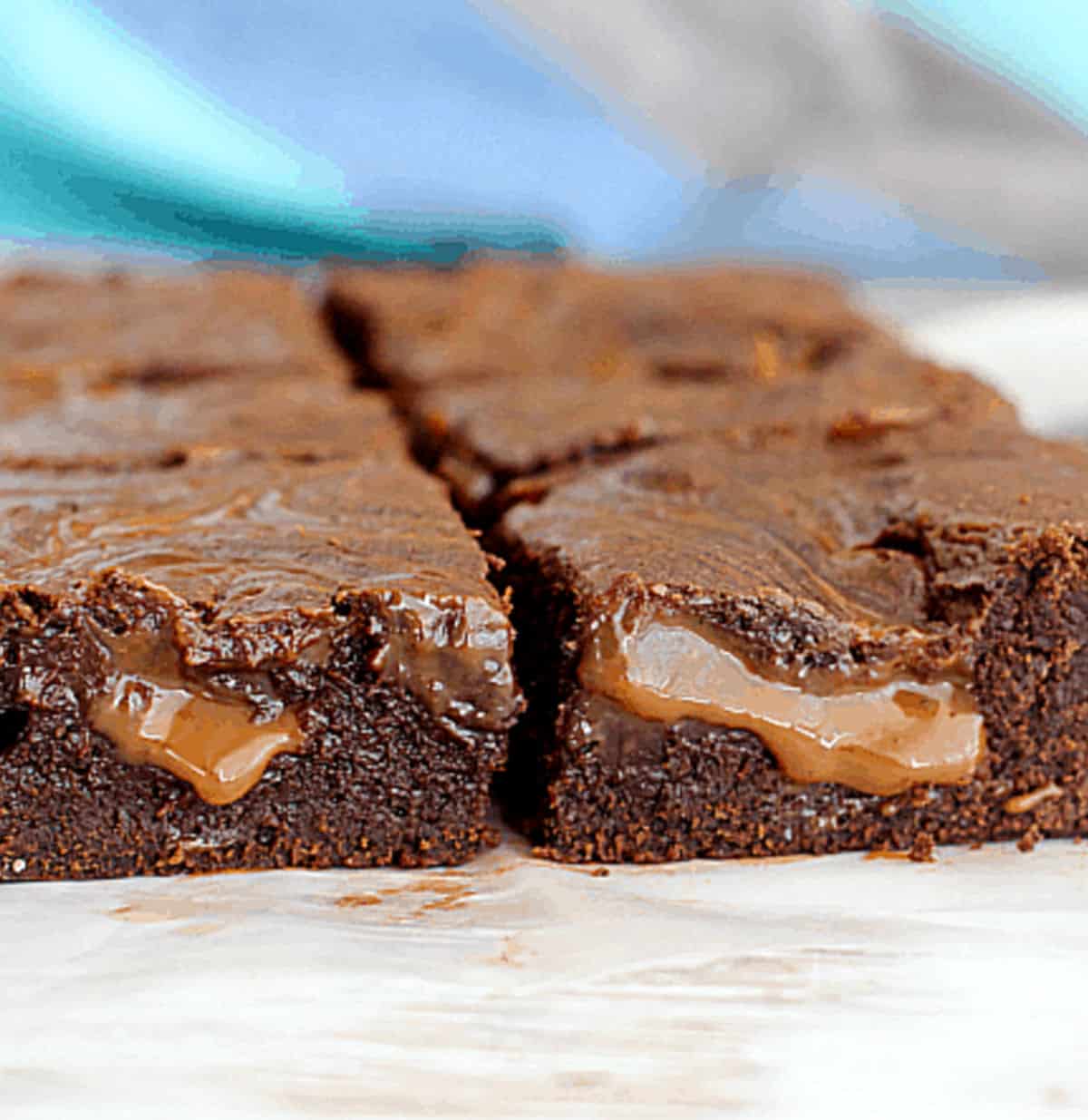 Squares of brownies filled with soft dulce de leche on white surface and bluish background.