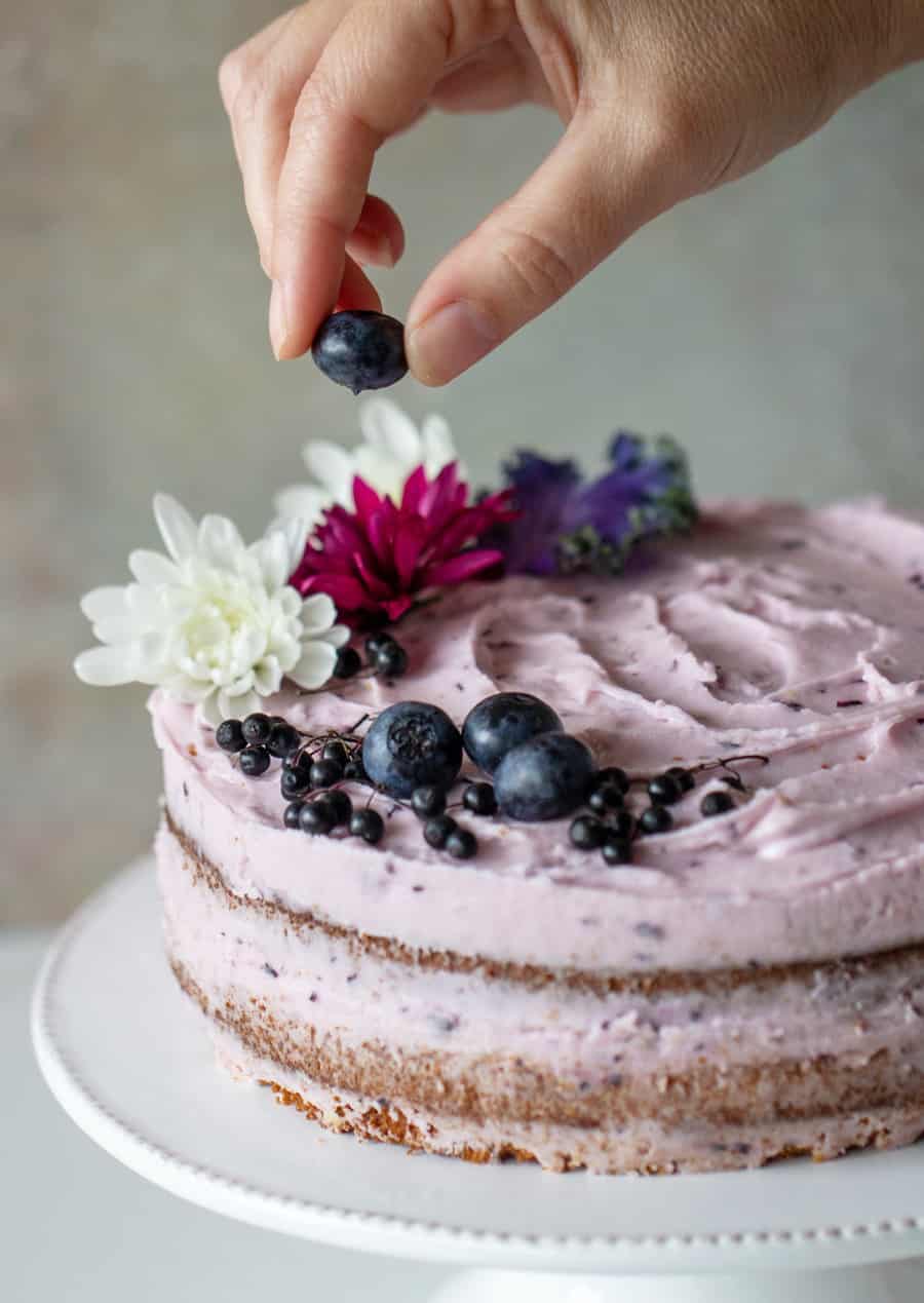 Whole decorated blueberry cake on a white cake stand, hand adding a blueberry.