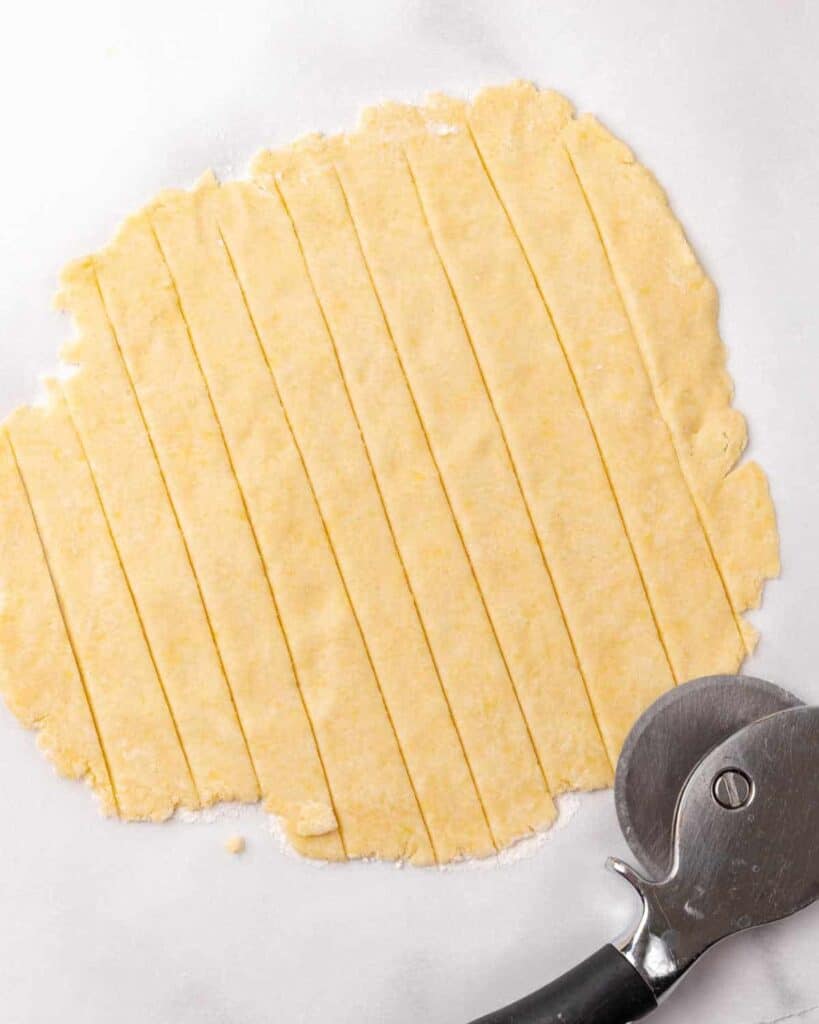 Strips cut on a rolled piece of pie dough on white marble. Pizza cutter.