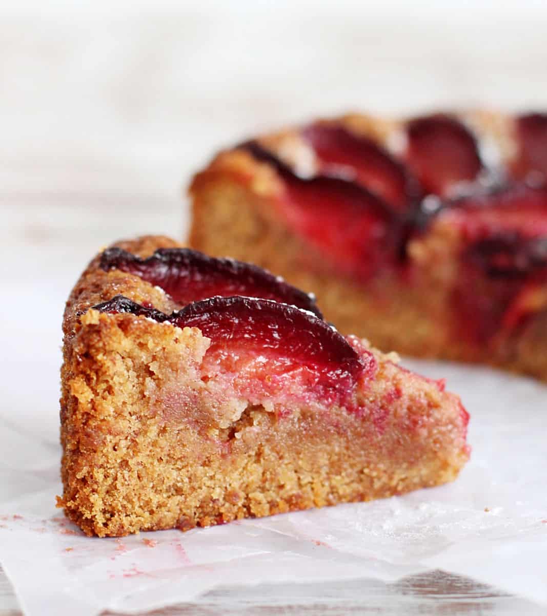 Slice of almond plum cake on white surface, rest of cake in background.