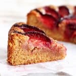 Single slice of honey almond plum cake on a piece of white paper. Rest of cake in the background.