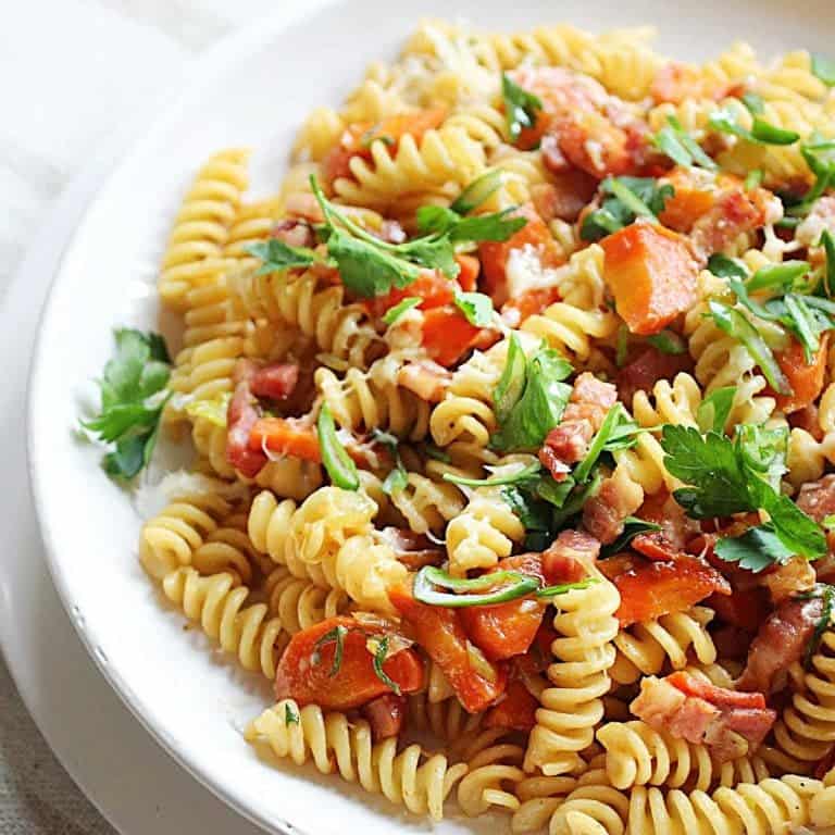 White plate with fusilli pasta, carrots, bacon and chopped parsley.