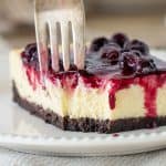 Close-up of cheesecake square on white plate, fork digging into it, berry topping