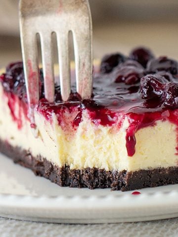 Close-up of cheesecake square on white plate, fork digging into it, berry topping