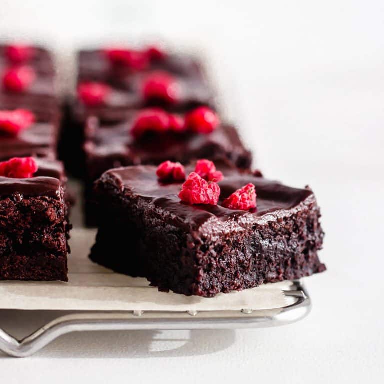 Squares of cocoa brownies, raspberry pieces, wire rack, white surface