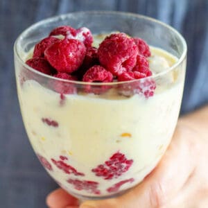 Hand holding a glass with lemon raspberry dessert, blue background.