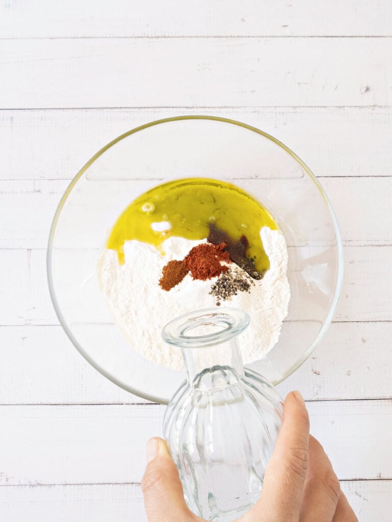 Glass bowl with ingredients for olive oil crackers. Hand adding water. White surface.