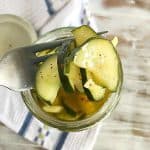 Close up of silver fork wit pickled cucumbers from a jar. White blue kitchen towel beneath on a washed white table.