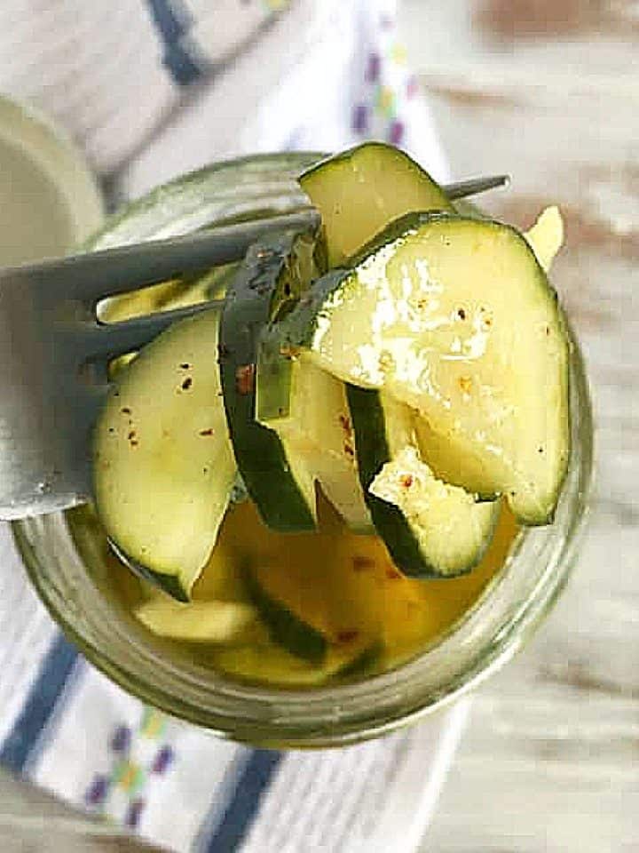 Close up of silver fork wit pickled cucumbers from a jar. White blue kitchen towel beneath on a washed white table.