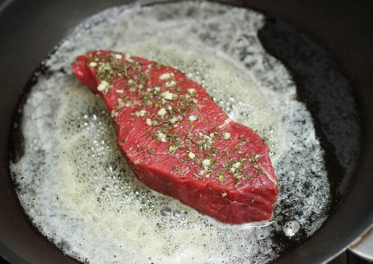 Raw seasoned rump steak in foamy butter on dark skillet