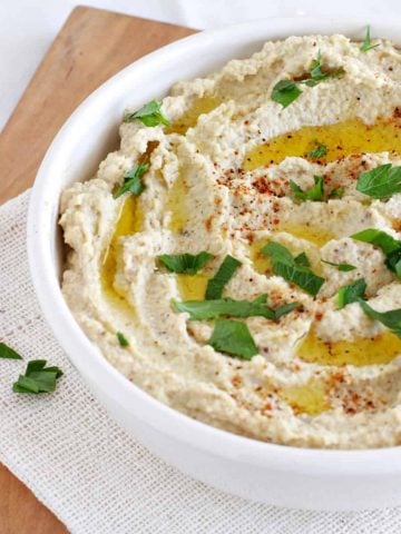 White bowl with eggplant hummus on wooden board. Parsley and olive oil garnish.