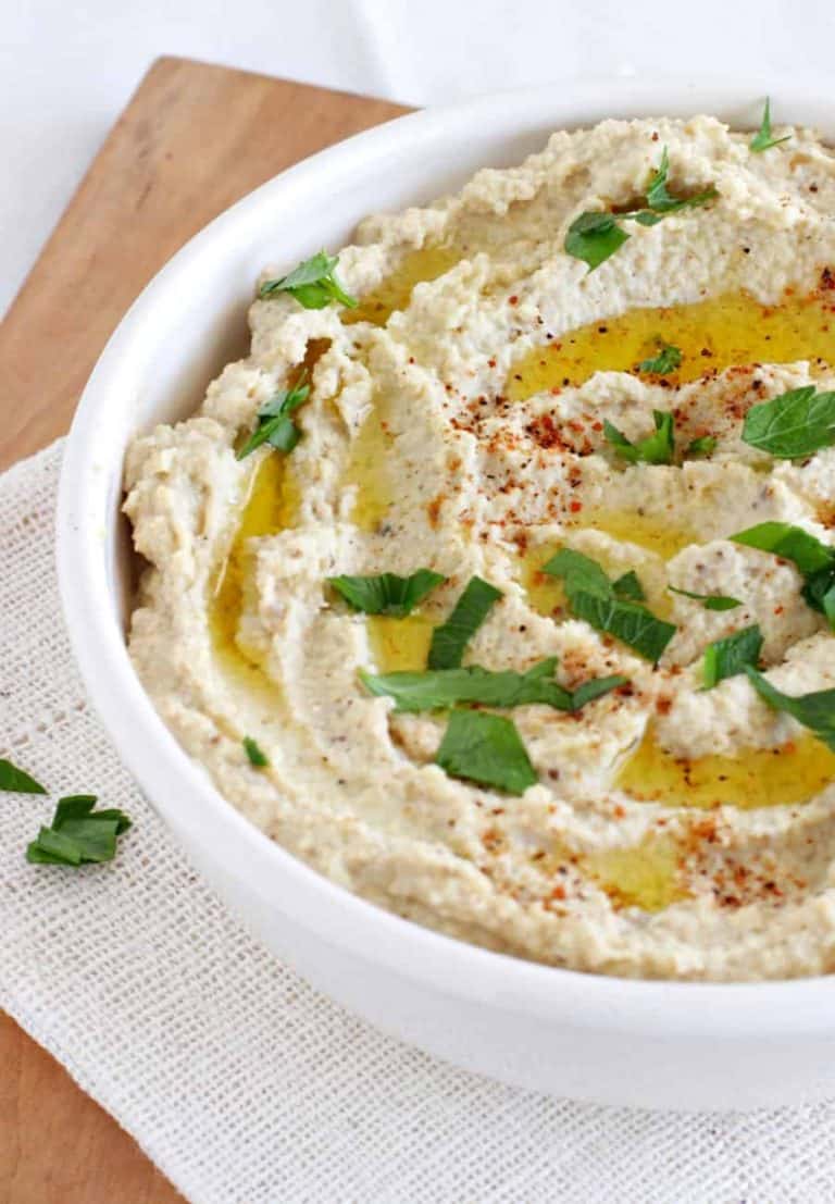 White bowl with eggplant hummus on wooden board. Parsley and olive oil garnish.