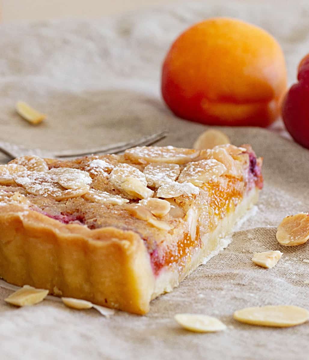 Slice of apricot tart on beige cloth, whole apricots, loose almonds.