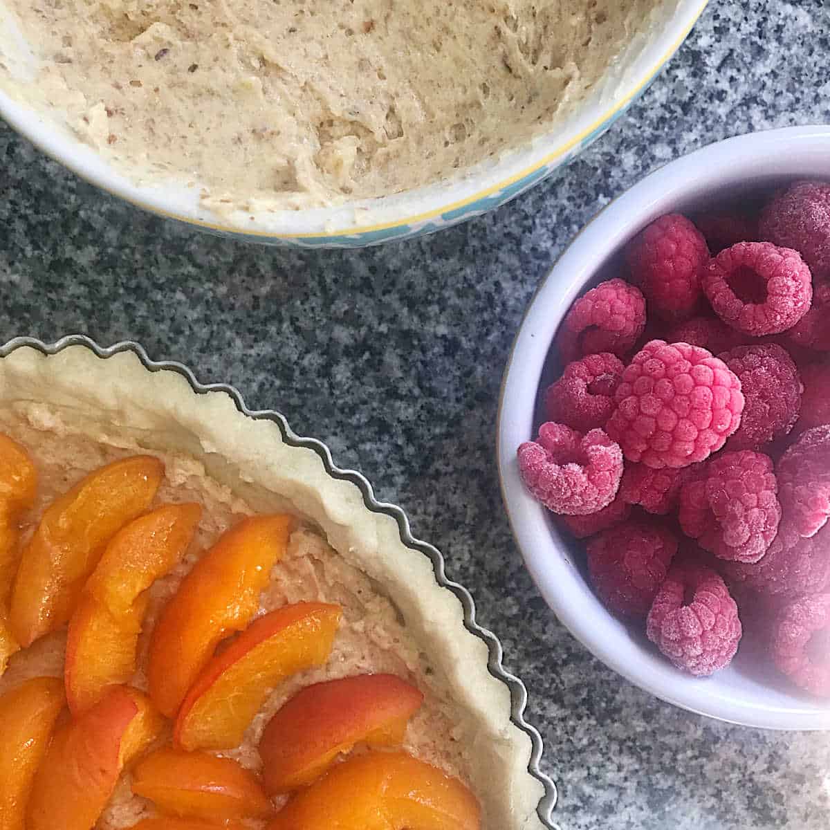 Partial view of crust with apricots, bowl with frangipane, raspberries in bowl.