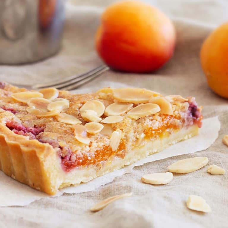 Slice of apricot frangipane tart on a beige cloth with sliced almonds and apricots around.