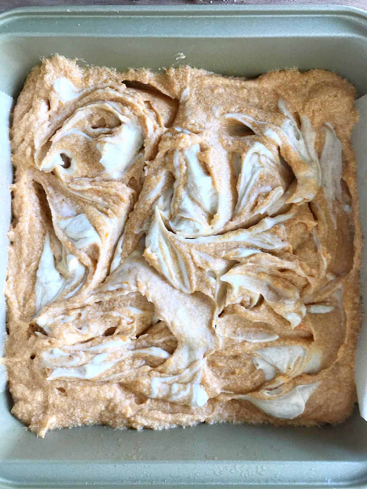 Swirled white mixture on pumpkin mixture in a metal square pan.