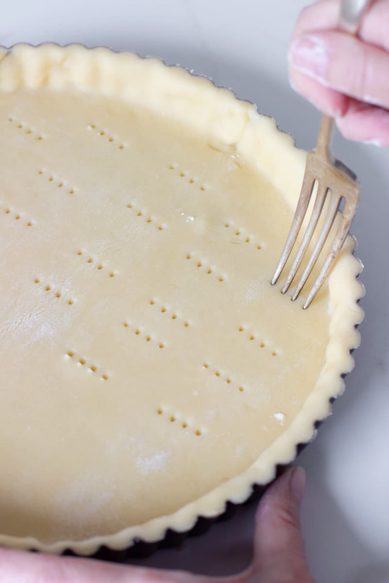 Fork pricking unbaked tart dough on white surface