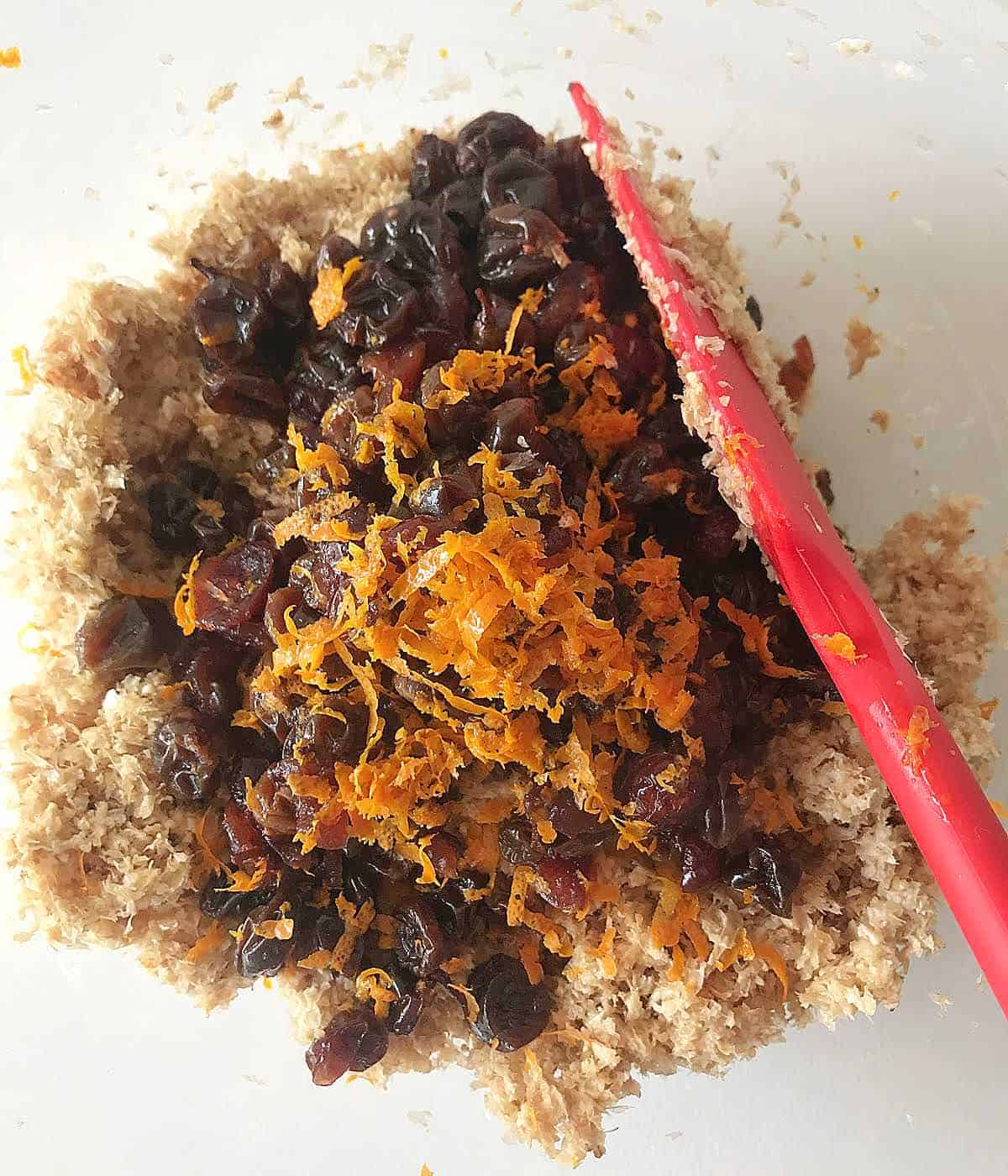 Raisins and orange peel added to bran muffin batter in a bowl with red spatula.
