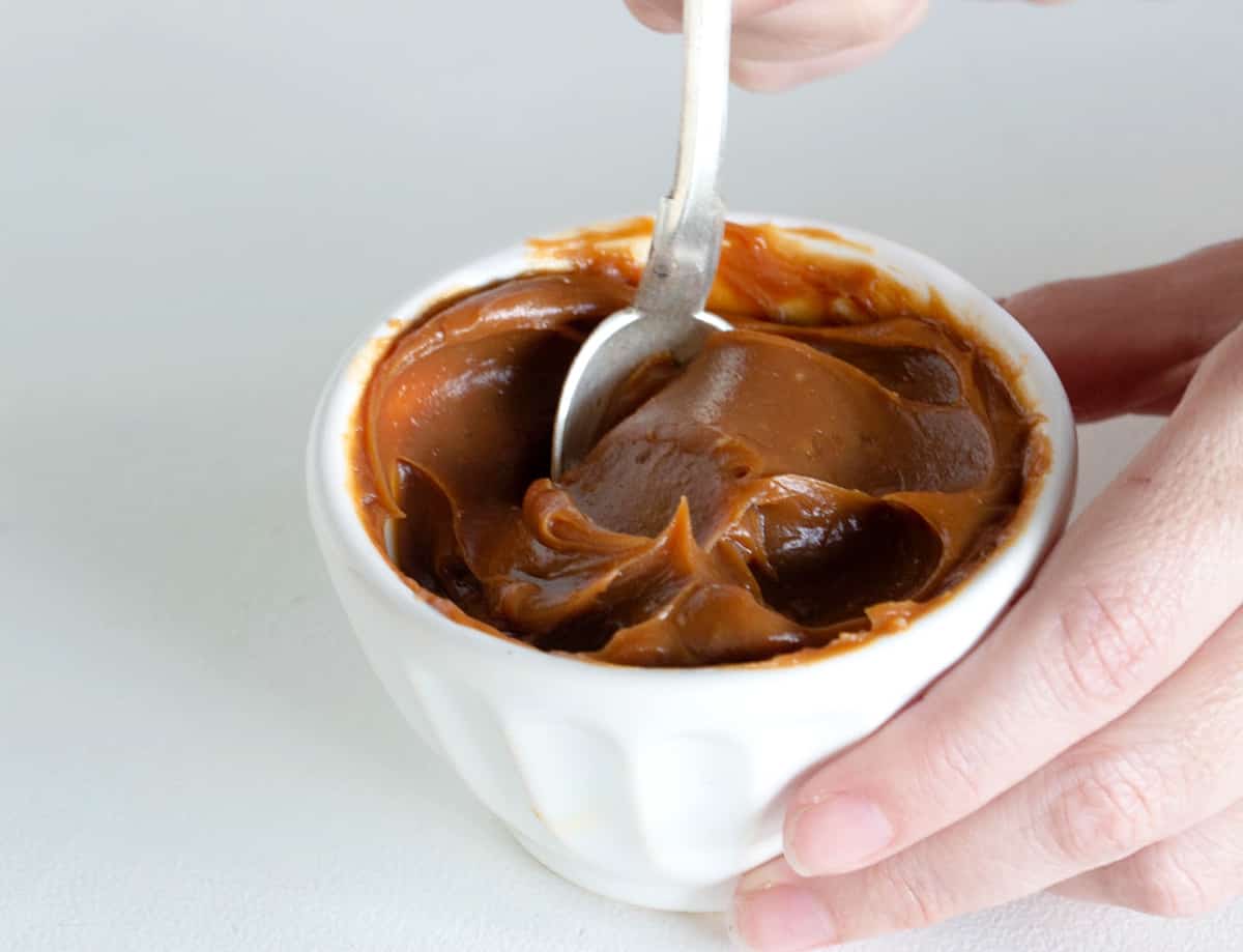 Hand dipping spoon in white bowl of dulce de leche