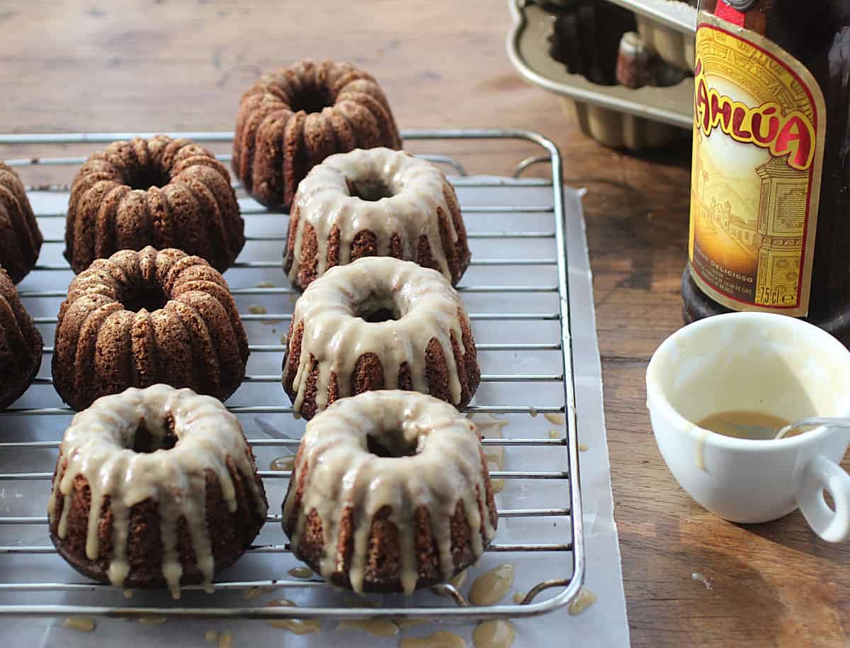 Bottle of kahlua, white cup with glaze, and mini chocolate bundt cakes on a wire rack with parchment paper on a wooden table. 
