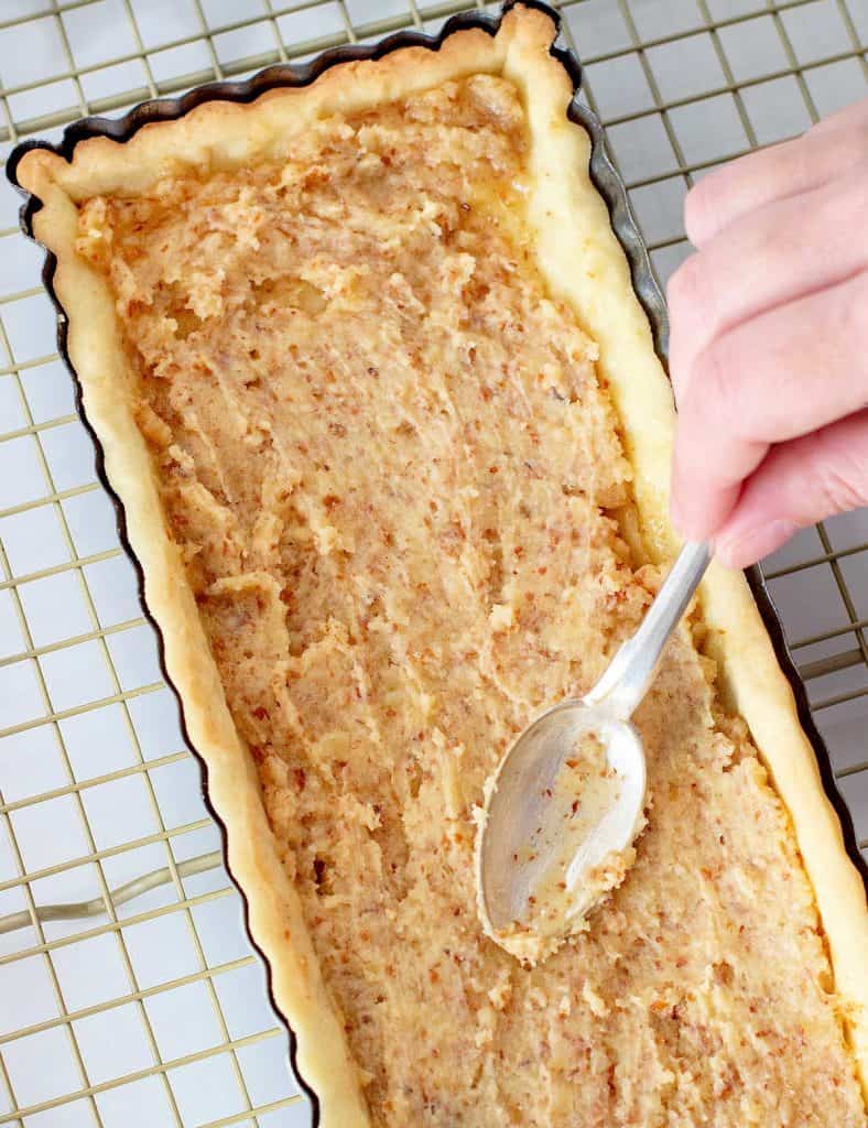 Hand spreading almond cream on rectangular pie crust on a wire rack.